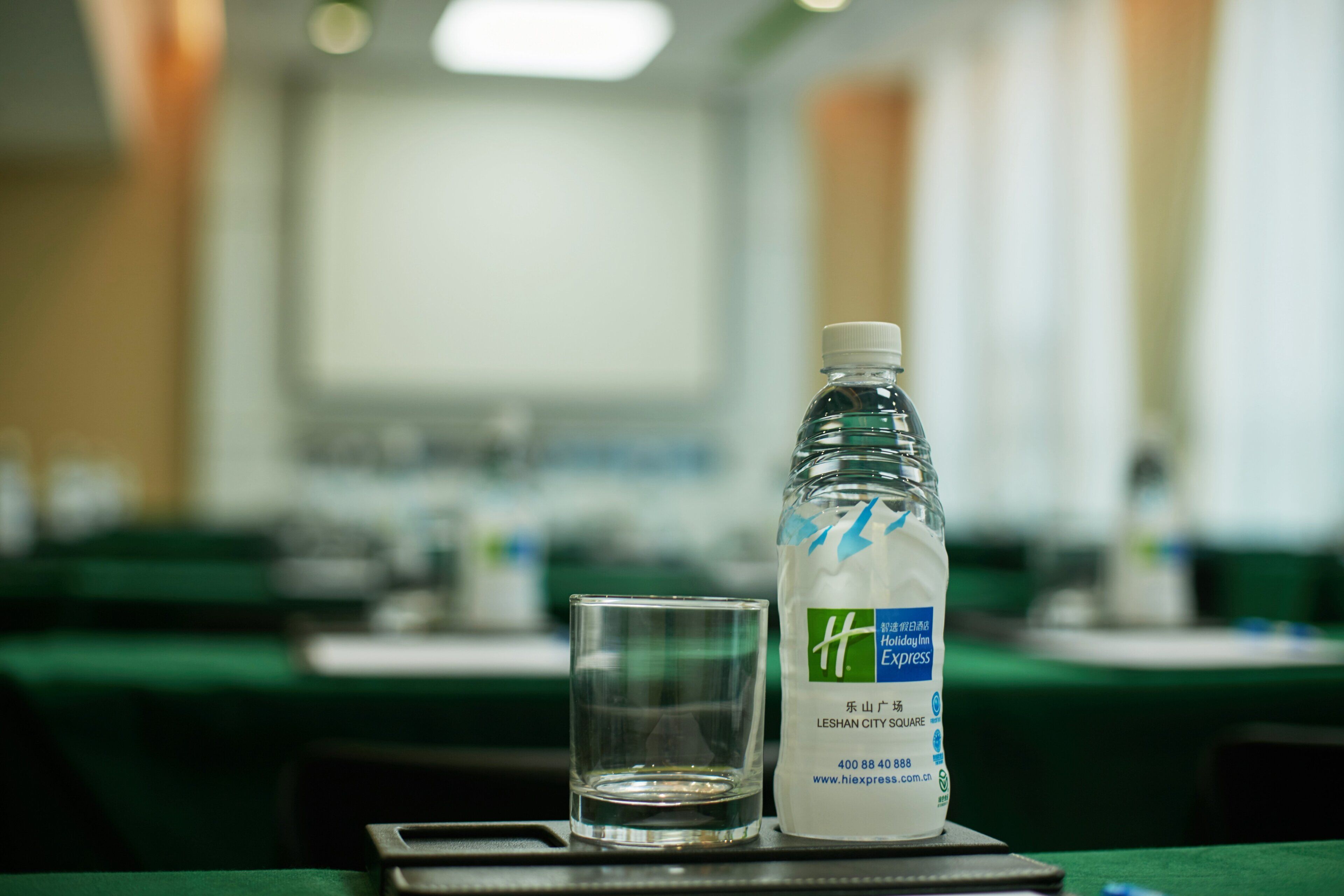 Une bouteille d’eau et un verre sur un plateau, dans une salle de conférence avec des tables et des chaises.