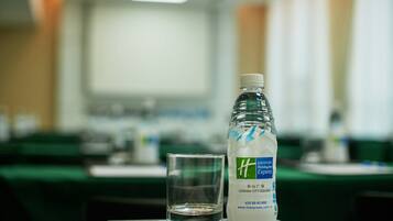 Une bouteille d’eau et un verre sur un plateau, dans une salle de conférence avec des tables et des chaises.
