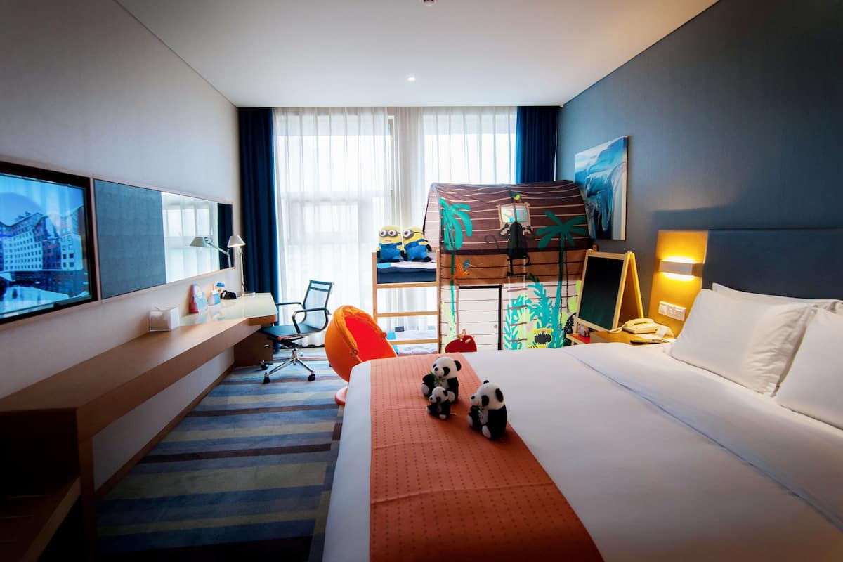 A hotel room with a large bed, a desk, a chair, a TV, and a bunk bed.