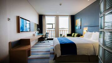 A modern hotel room with a large bed, a desk, a chair, and a view of the city through large windows.