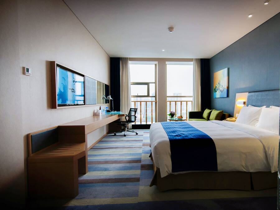 A modern hotel room with a large bed, a desk, a chair, and a view of the city through large windows.