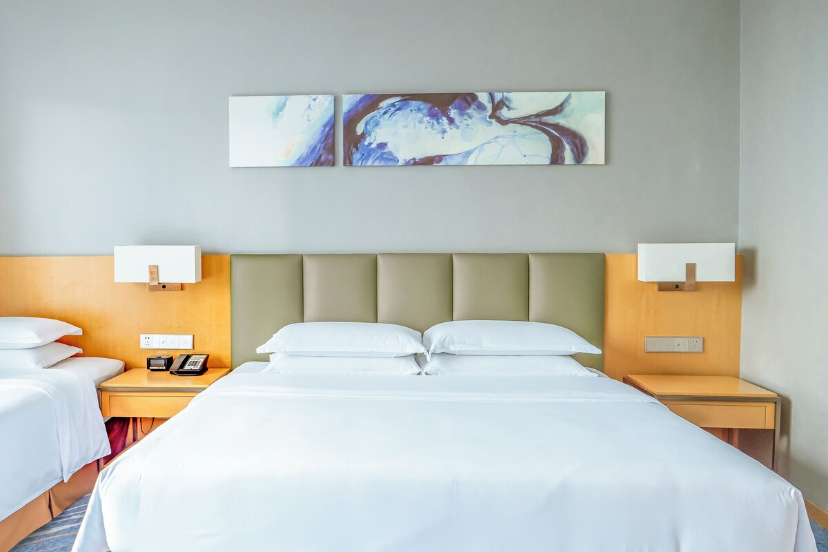 A hotel room with two single beds, a large bed, a headboard with integrated lighting, a bedside table with a phone, and a wall-mounted abstract art piece.