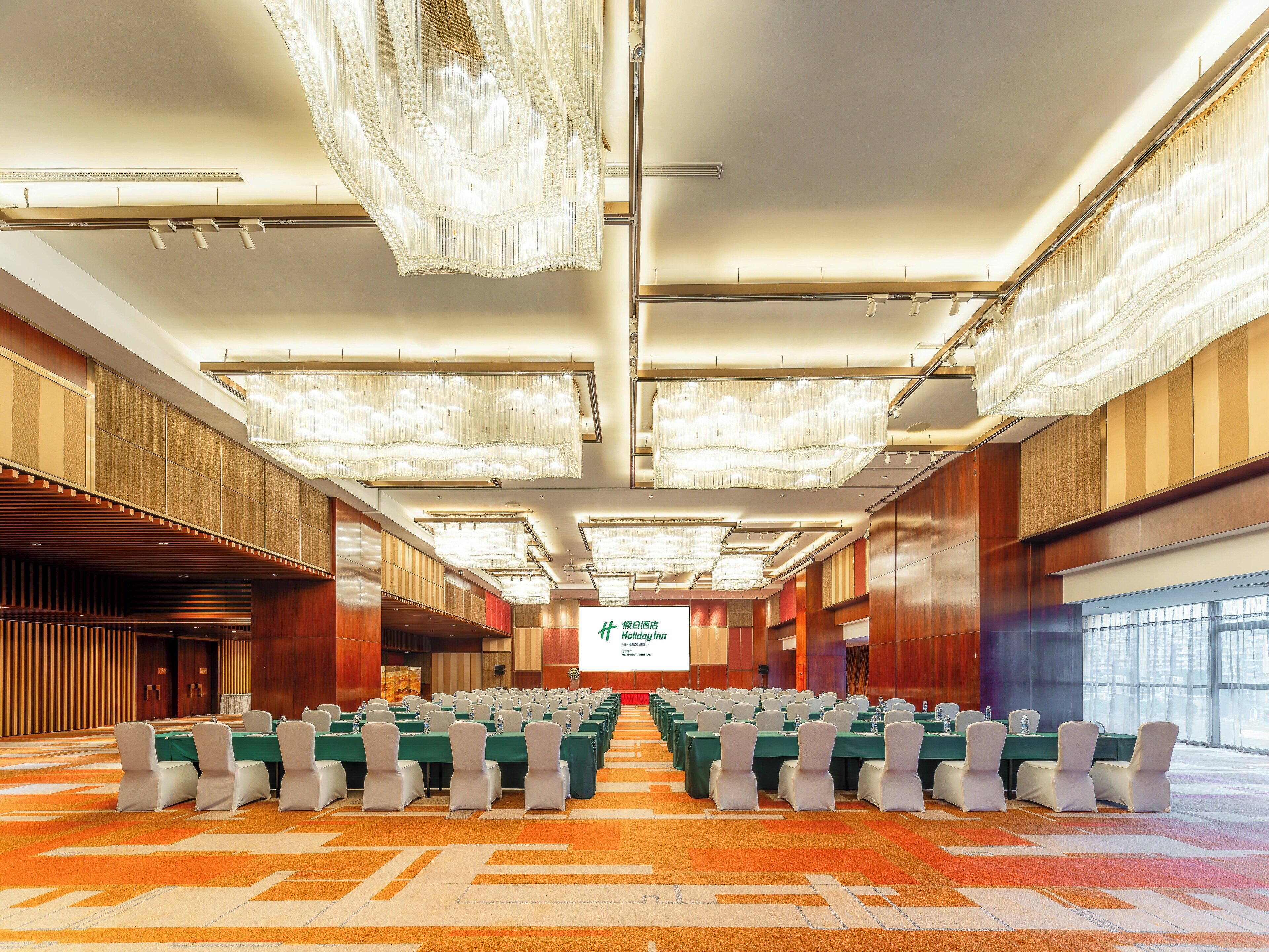 Un salón de conferencias amplio con filas de mesas y sillas, techos altos con candelabros y una pantalla grande al frente.