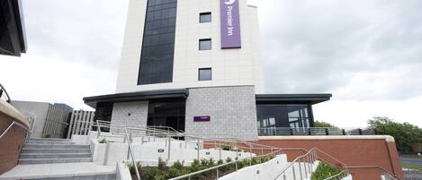A hotel entrance with a modern design, featuring a prominent 'Premier Inn' sign, a set of stairs leading to the entrance, and a clear sky above.