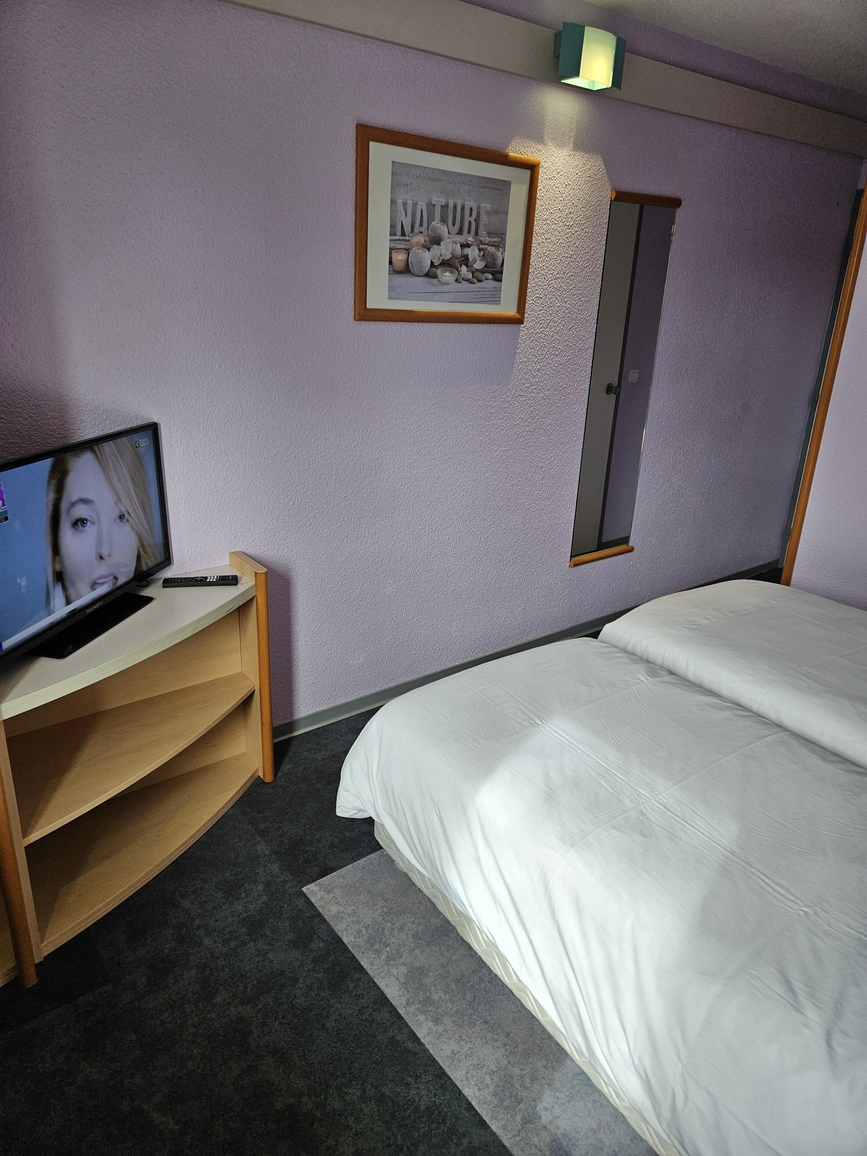 A hotel room with a bed, a desk with a computer, a framed picture on the wall, and a door.