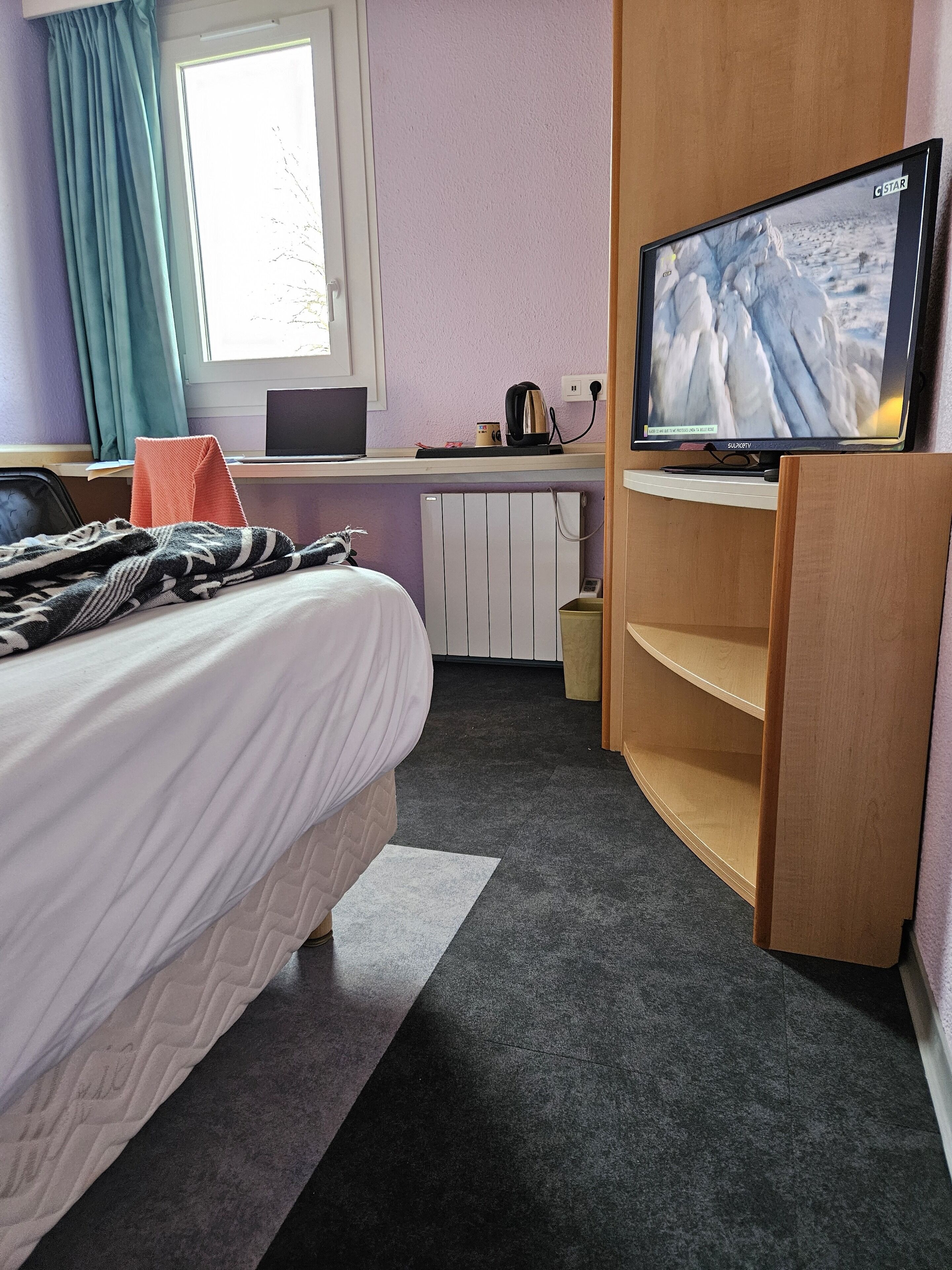 Quarto de hotel com cama, secretária com laptop, televisão e radiador.