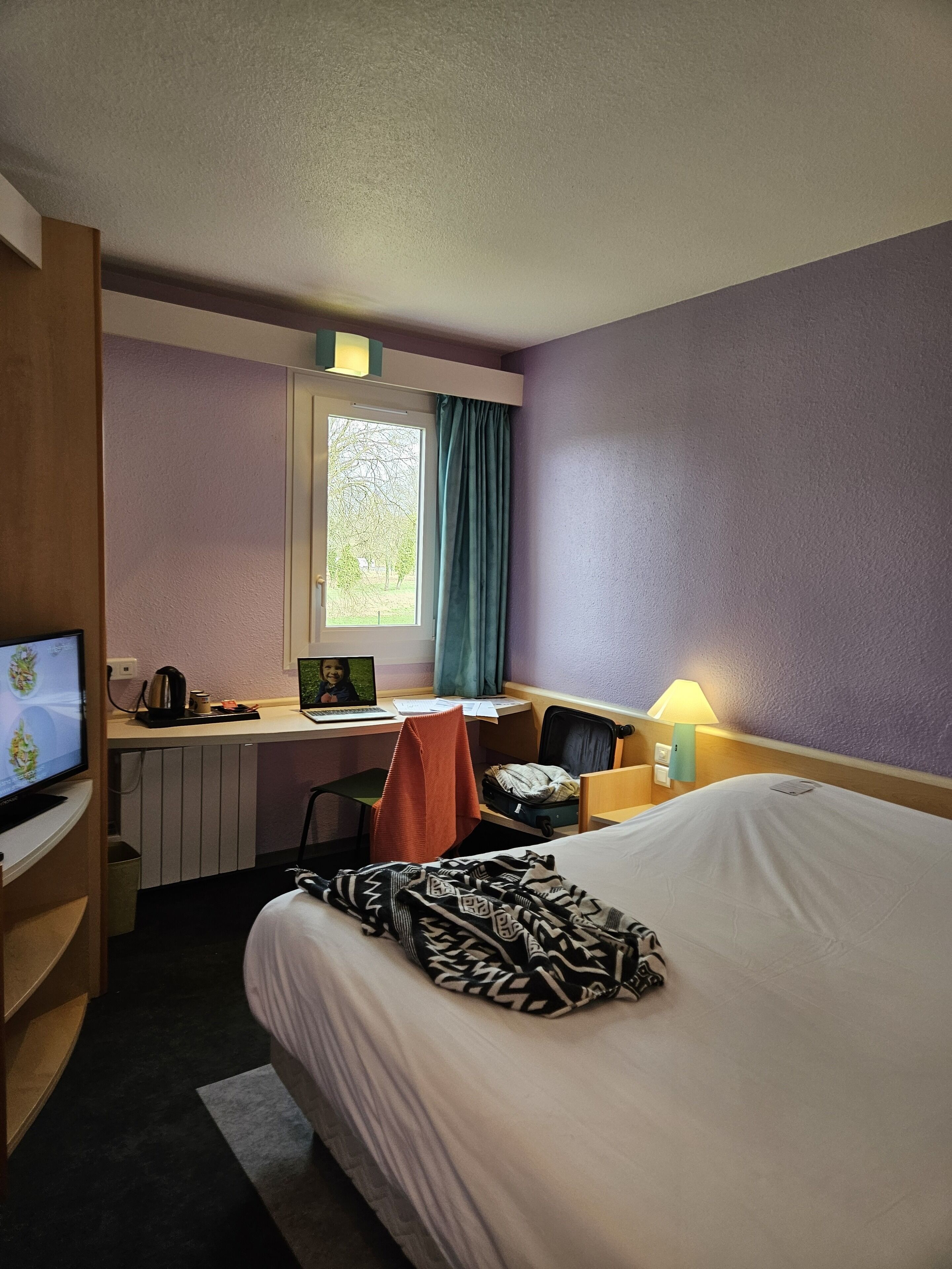 Habitación de hotel con cama, escritorio con una computadora portátil, silla, televisor y ventana con cortinas.