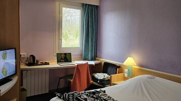 A hotel room with a bed, a desk with a laptop, a chair, a television, and a window with curtains.