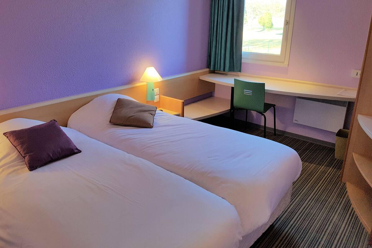 A hotel room with two beds, a desk, a chair, a lamp, and a window with curtains.