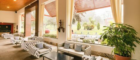 A spacious indoor seating area with multiple sofas and a coffee table, large windows with blinds, and a potted plant.