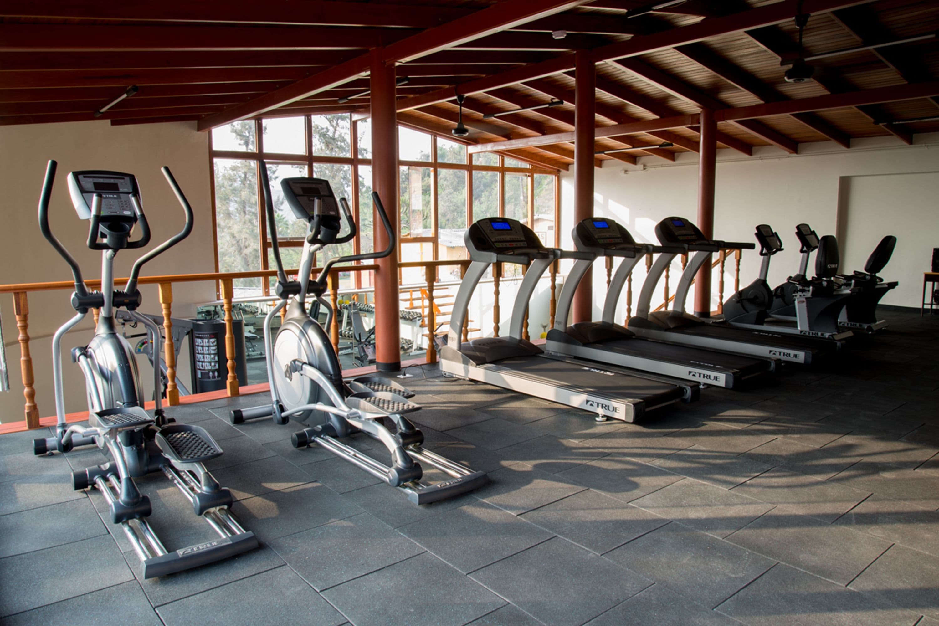A gym with various cardio machines, including ellipticals and treadmills, under a wooden roof with large windows.