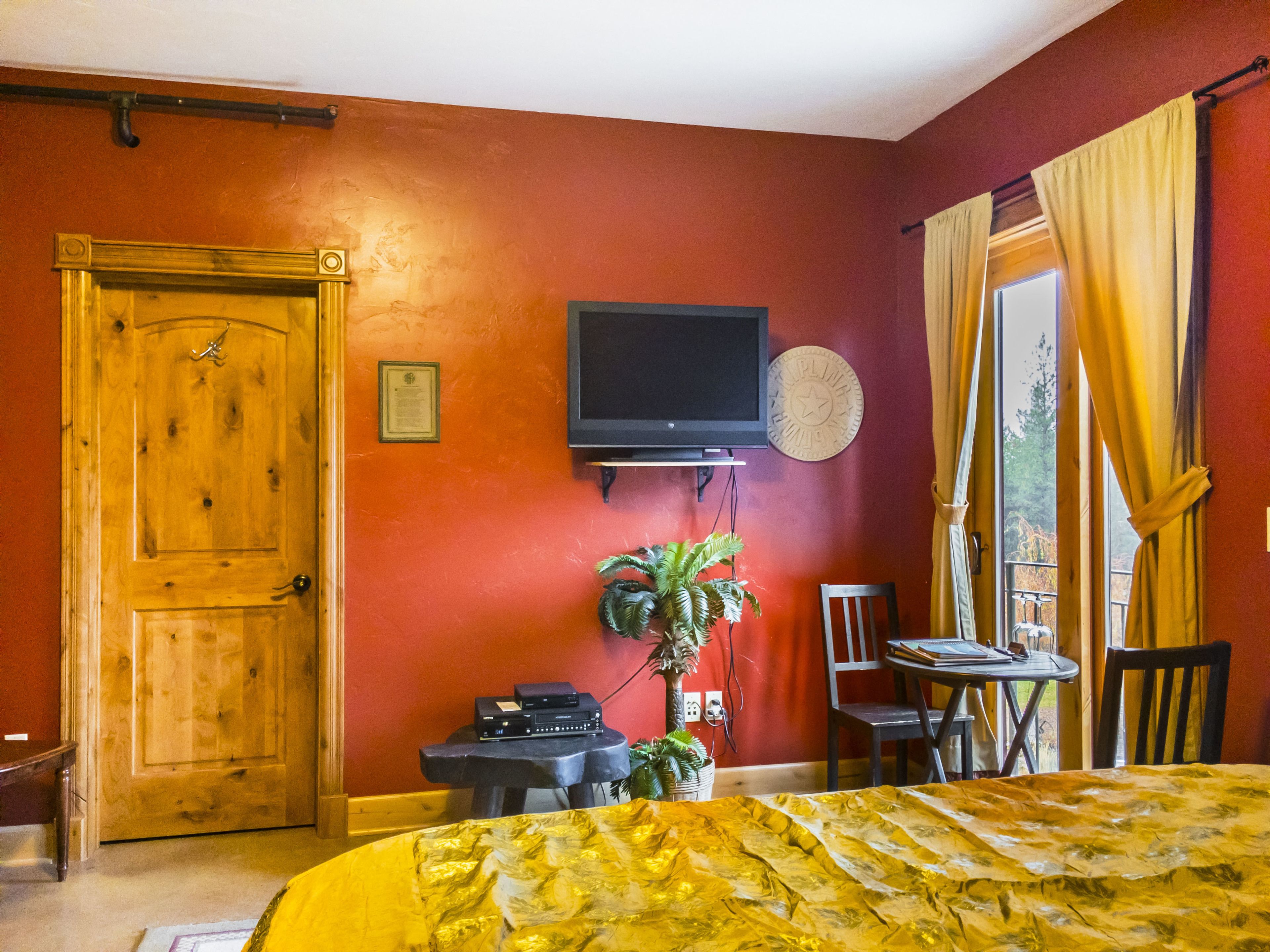 A room with a wooden door, a television mounted on the wall, a small table with a chair, and a window with curtains.