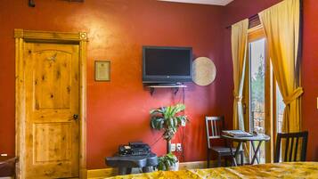 A room with a wooden door, a television mounted on the wall, a small table with a chair, and a window with curtains.