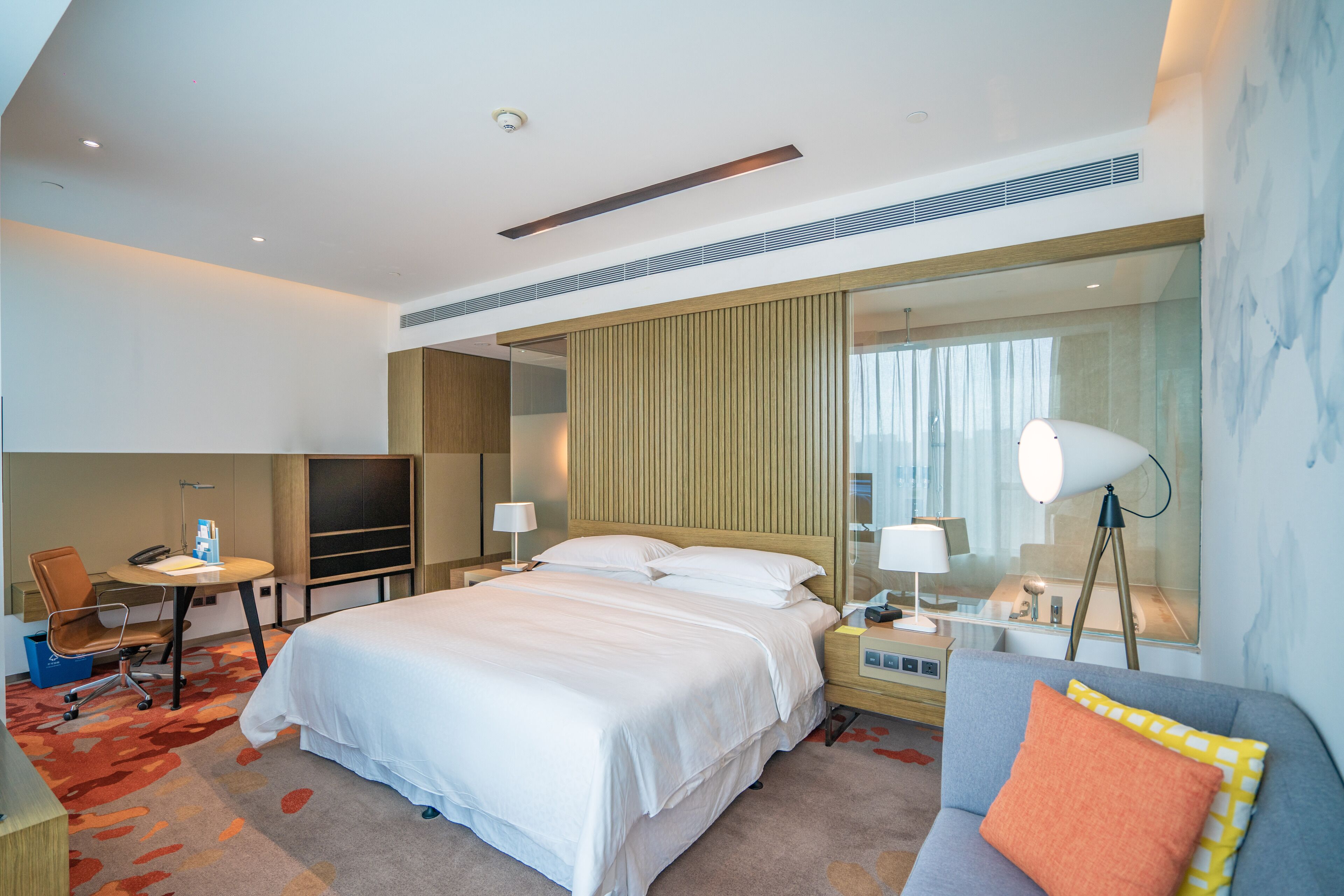 A modern hotel room with a large bed, a desk, a chair, a TV, a lamp, and a sofa.