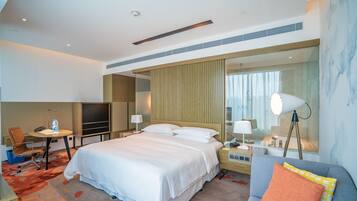 A modern hotel room with a large bed, a desk, a chair, a TV, a lamp, and a sofa.