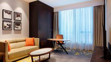 A modern hotel room with a sofa, a small round table, a chair, a TV, and a large window with curtains.