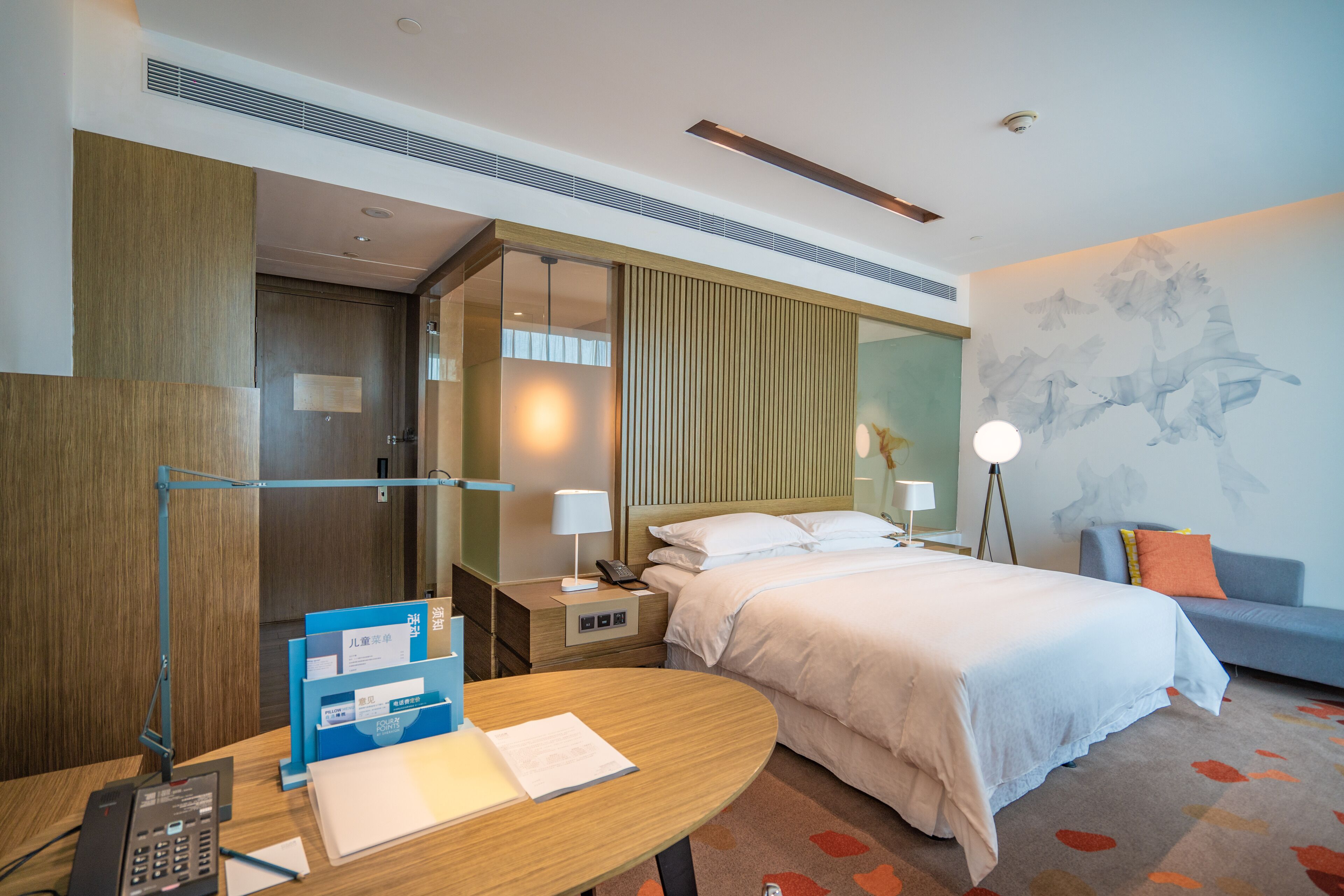 A hotel room with a bed, bedside table, lamp, and a desk with a telephone and papers.