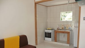 A compact living space with a kitchenette, a sofa with a yellow throw, and a window overlooking a garden. - Morada das Palmeiras (Maraú)
