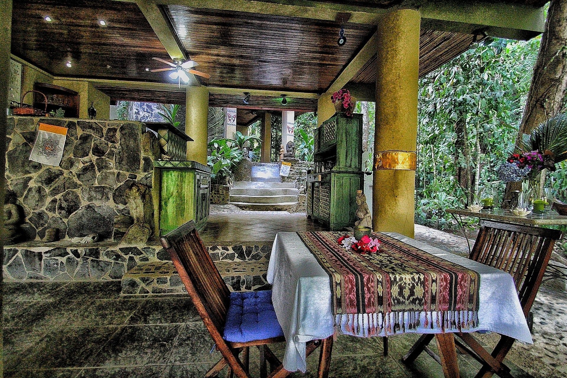 An outdoor covered dining area with stone walls, a table with a tablecloth, and chairs.