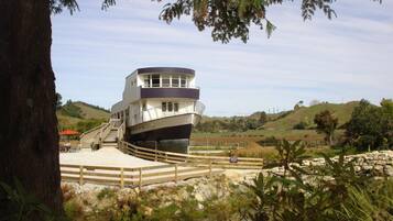 Un edificio con forma de barco, con una escalera que lleva a la cubierta superior, rodeado de paisaje natural y una valla de madera.