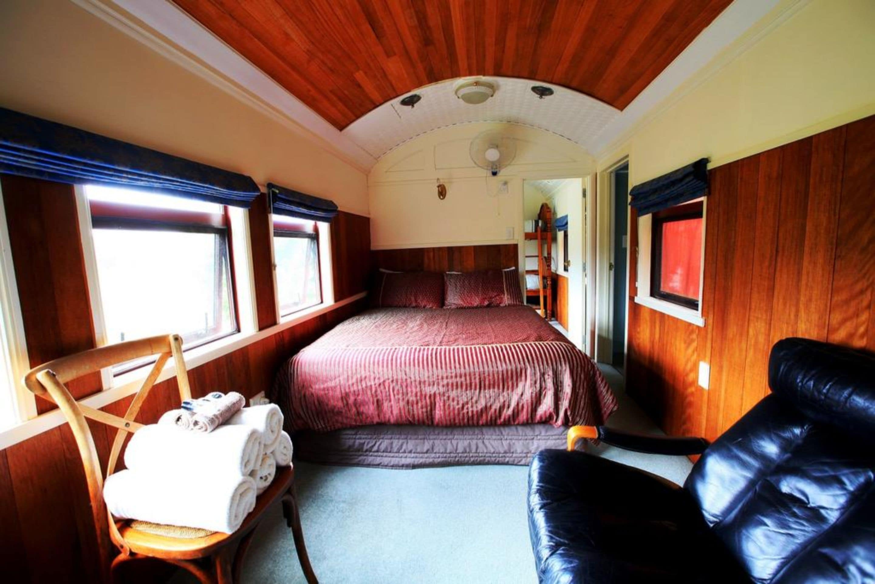 Un vagón de tren con interior de madera, una cama con ropa de cama roja, una silla de madera con toallas y un sofá de cuero negro.