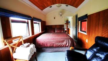 Un vagón de tren con interior de madera, una cama con ropa de cama roja, una silla de madera con toallas y un sofá de cuero negro.
