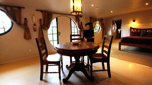 A dining area with a round wooden table and chairs, a hanging light fixture, and large windows with curtains. - Woodlyn Park (Waitomo Caves)