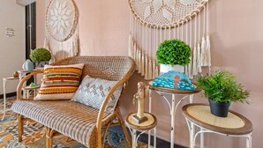 A wicker sofa with patterned cushions, a macrame wall hanging, a small round table with a potted plant, and a decorative statue.