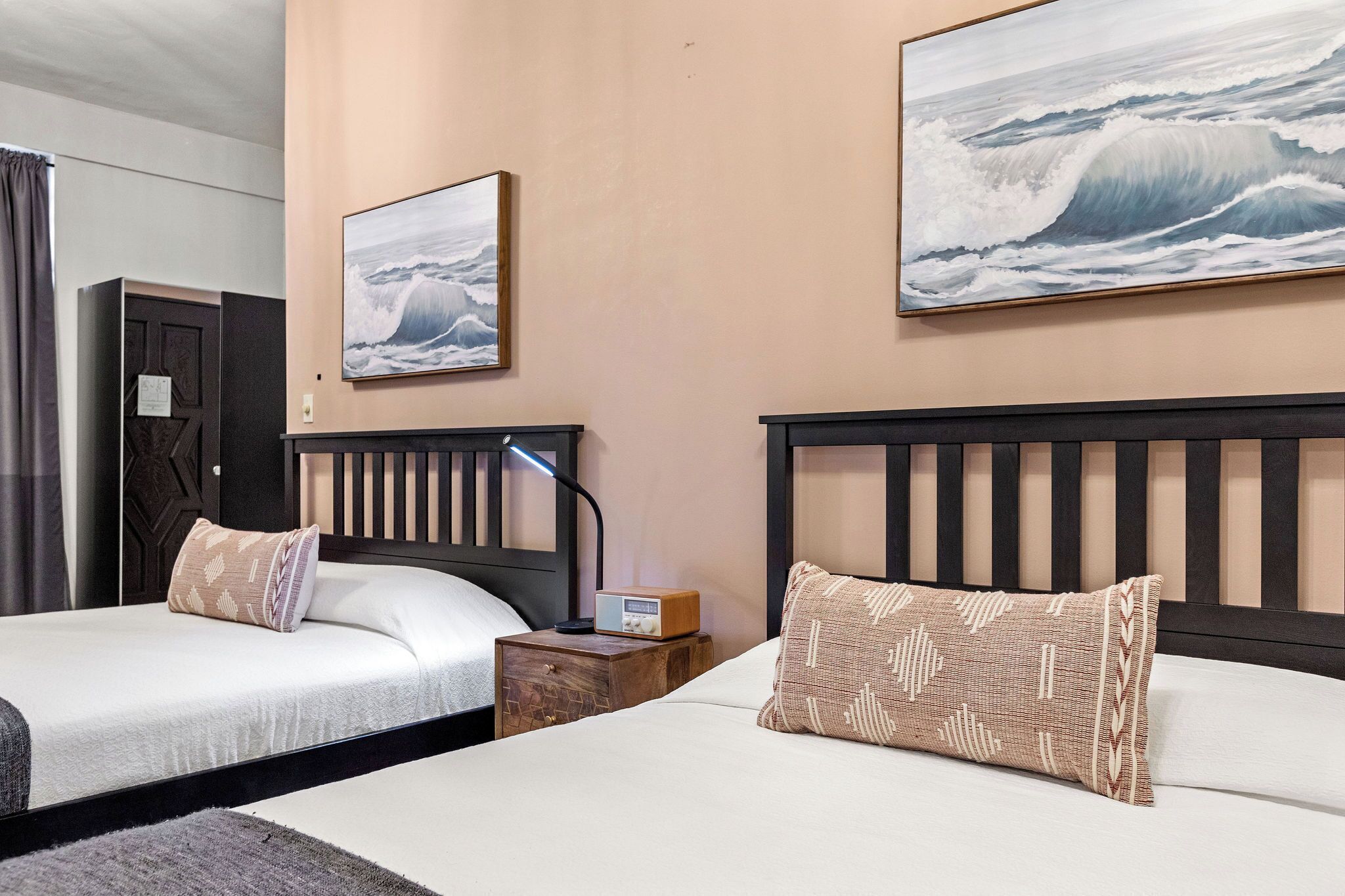 A hotel room with two beds, a nightstand with a radio, and two large ocean wave paintings on the walls.