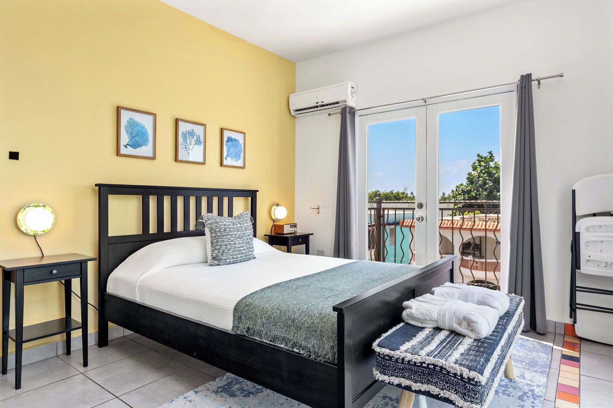 A bedroom with a bed, bedside tables, a bench, a balcony with a view, and an air conditioning unit.