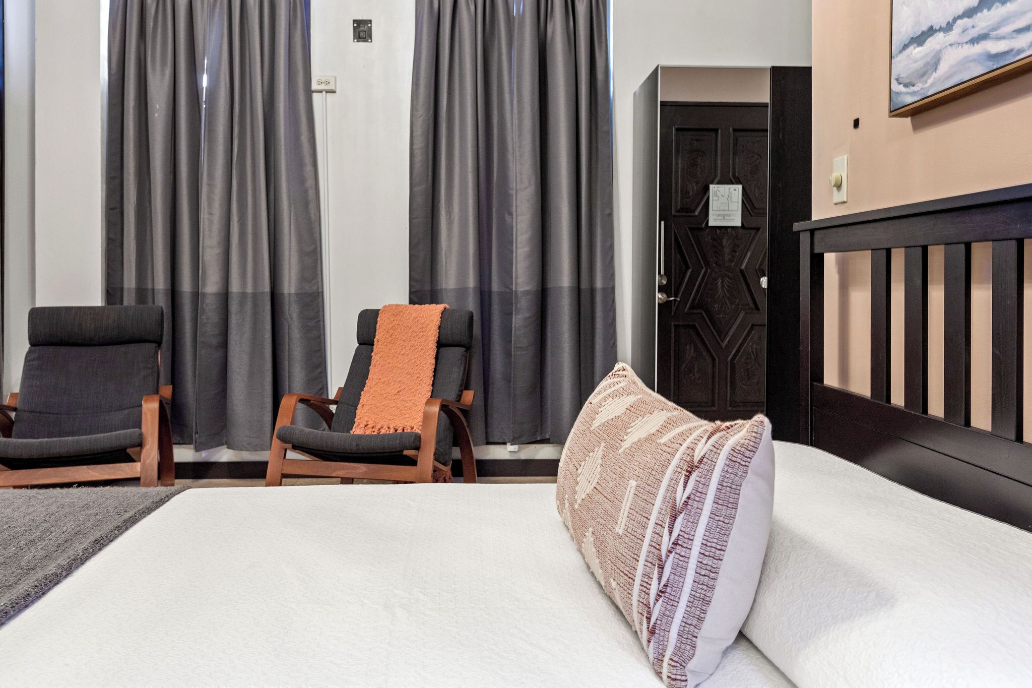 Ein Hotelzimmer mit einem Bett, zwei Sesseln, Blick nach draußen durch Vorhänge und einer Tür mit geometrischem Muster.