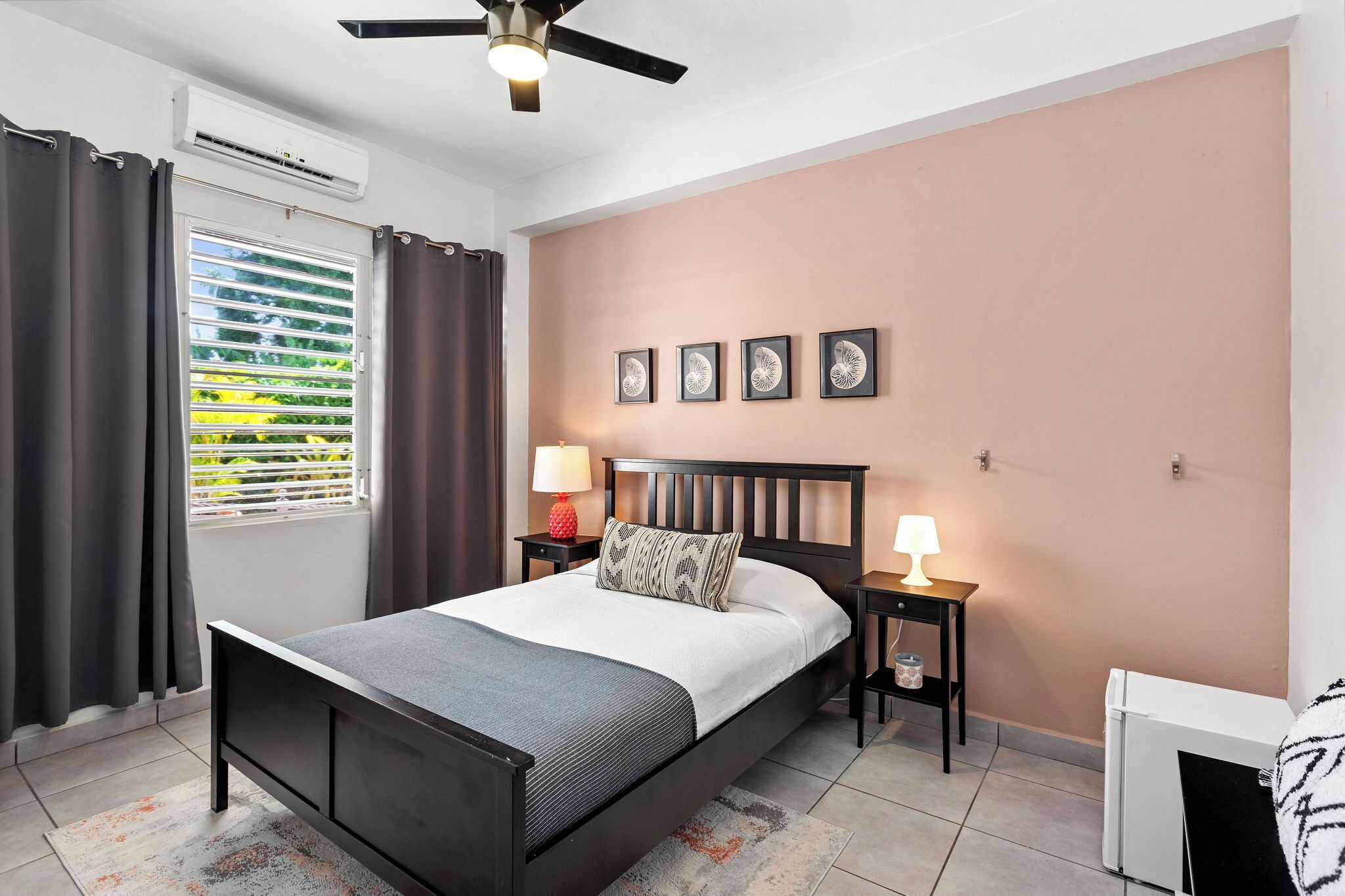 A bedroom with a bed, a ceiling fan, an air conditioning unit, a window with curtains, a wall-mounted lamp, and a radiator.