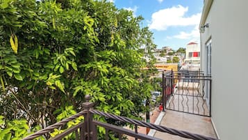 Un balcón con barandilla de metal que da a una calle con edificios y un árbol de hojas verdes.