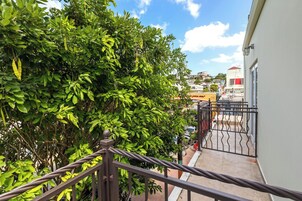 Un balcón con barandilla de metal que da a una calle con edificios y un árbol de hojas verdes.