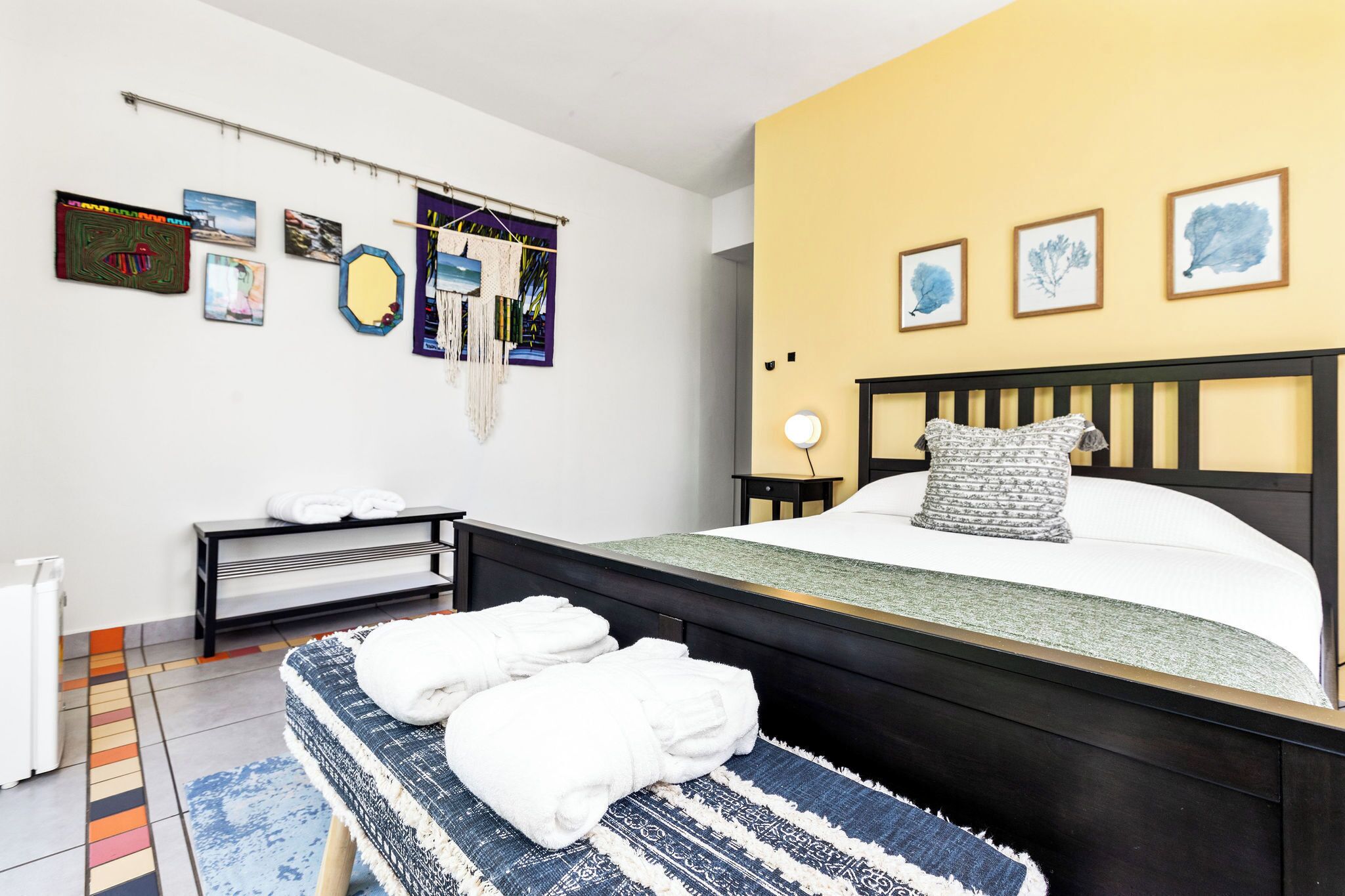 A hotel room with a bed, a bench with towels, a bedside table, and wall art.