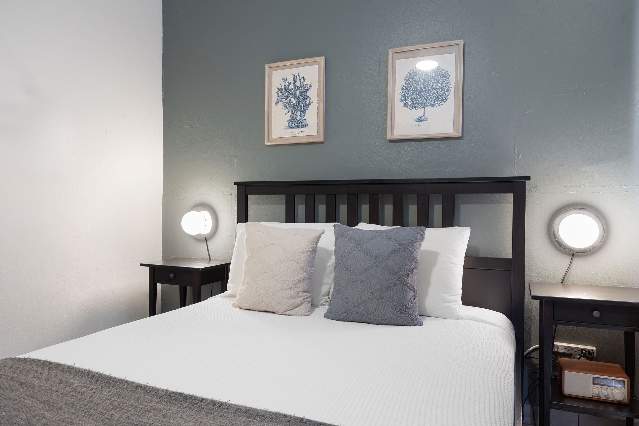 A neatly made bed with two pillows, a bedside table with a lamp, and two framed pictures on the wall.