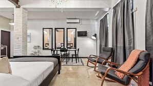 A modern hotel room with a bed, a desk, two chairs, a chandelier, and a large window with curtains.