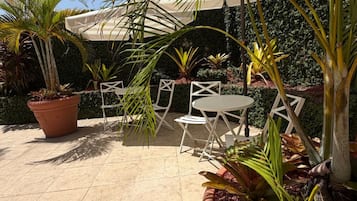 Un patio al aire libre con sillas blancas y una mesa, rodeado de plantas en macetas y un paraguas grande.