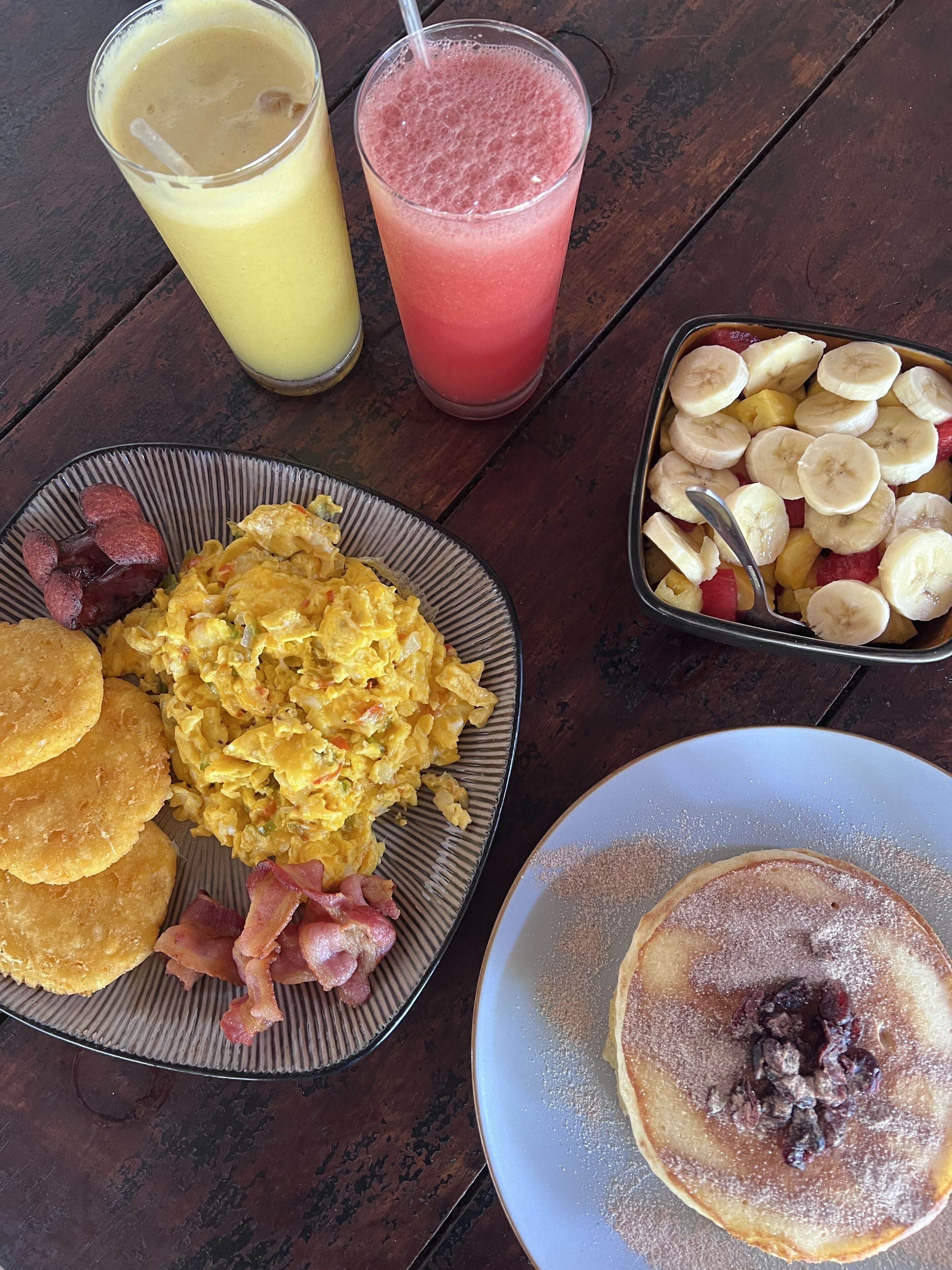 A breakfast plate with scrambled eggs, bacon, and fried plantains, a glass of orange juice, a glass of pink smoothie, and a plate of sliced bananas and pineapple.