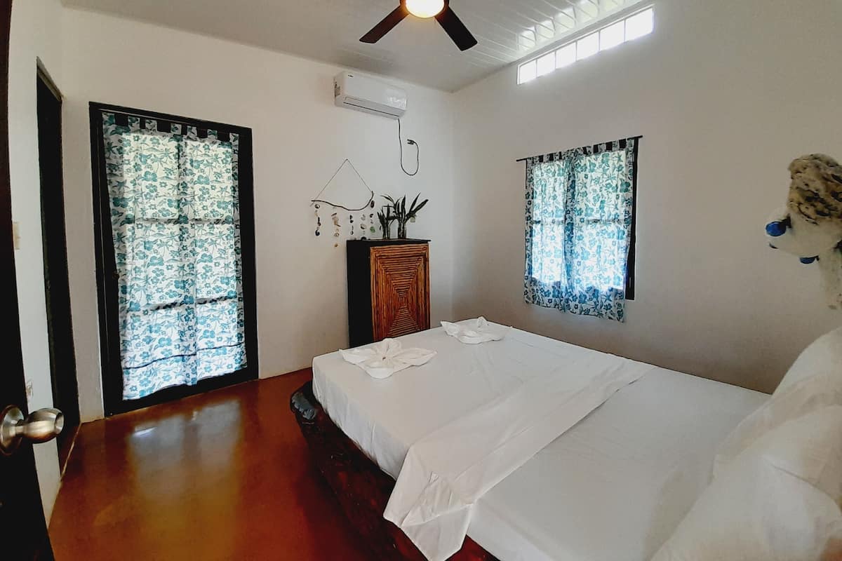 A bedroom with a bed, a ceiling fan, an air conditioning unit, and windows with curtains.