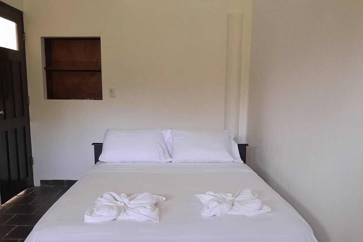 A neatly made bed with white linens and towels, a wooden door, and a small window with a wooden frame.