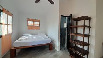 Un dormitorio con cama, ventilador de techo, estante de madera y baño visible a través de una puerta abierta.