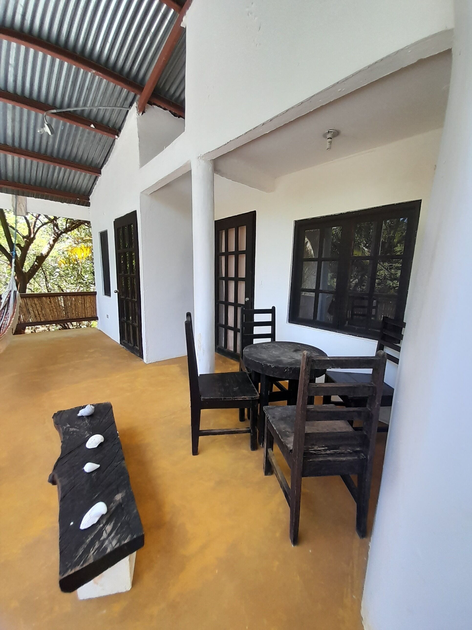 Un patio techado con una mesa, sillas, un banco y una ventana con vistas a árboles.