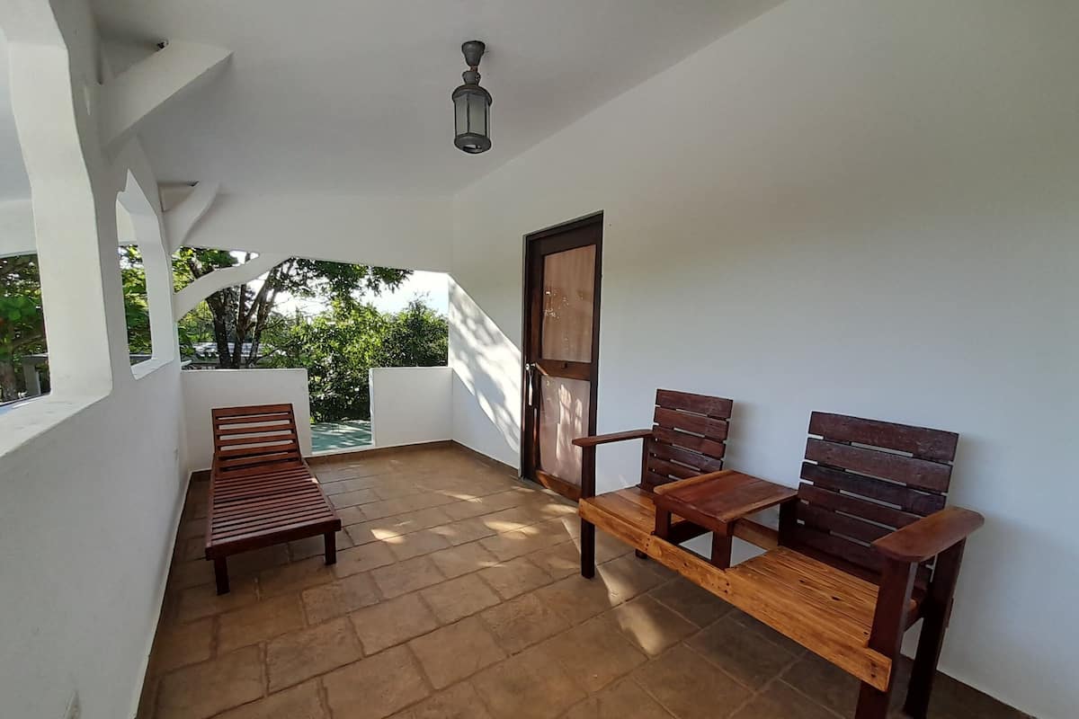 A balcony with a wooden bench and two chairs, a door, and a view of greenery.