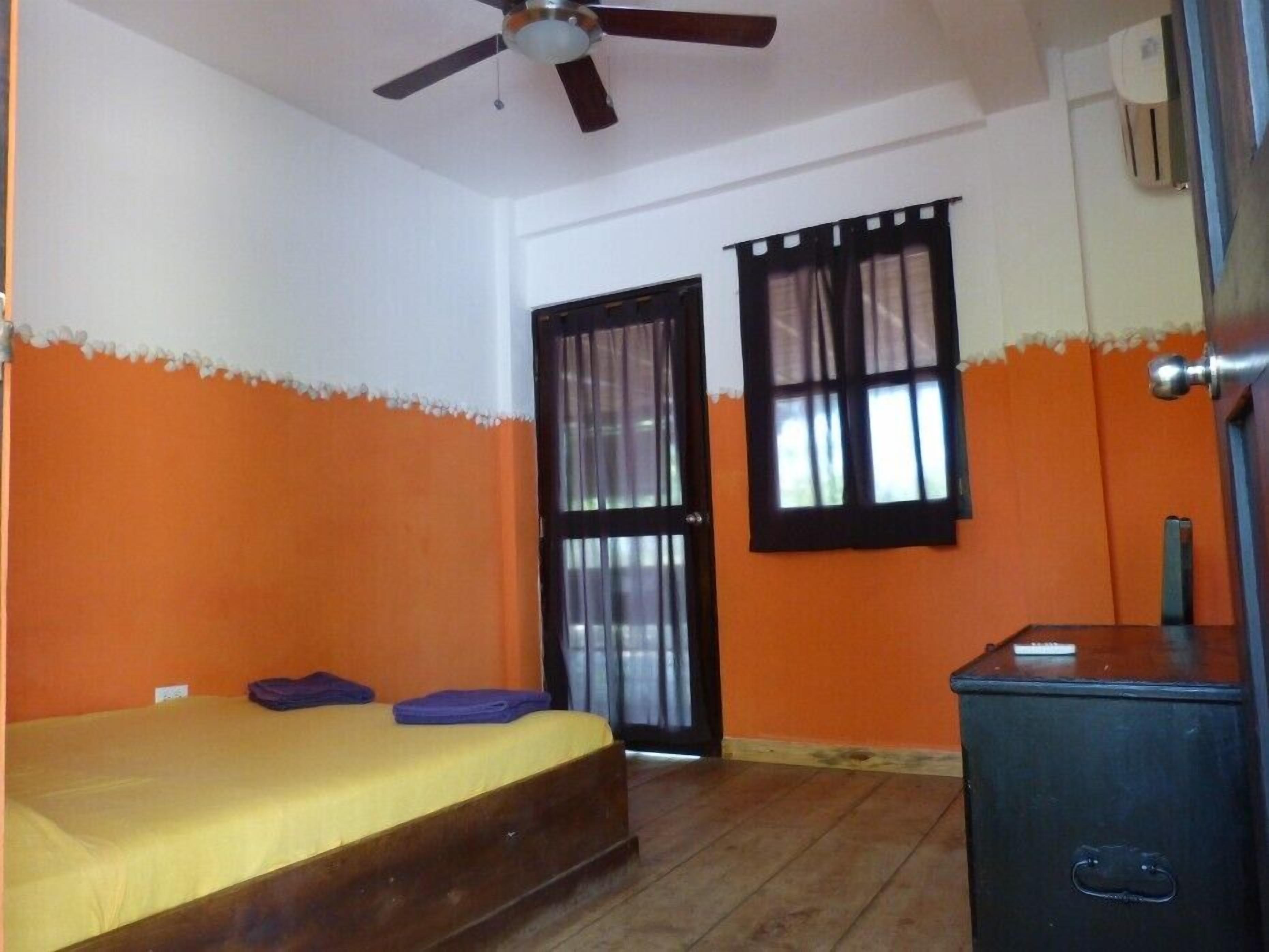 Une pièce avec un lit, un ventilateur de plafond, une fenêtre avec des rideaux et une porte.