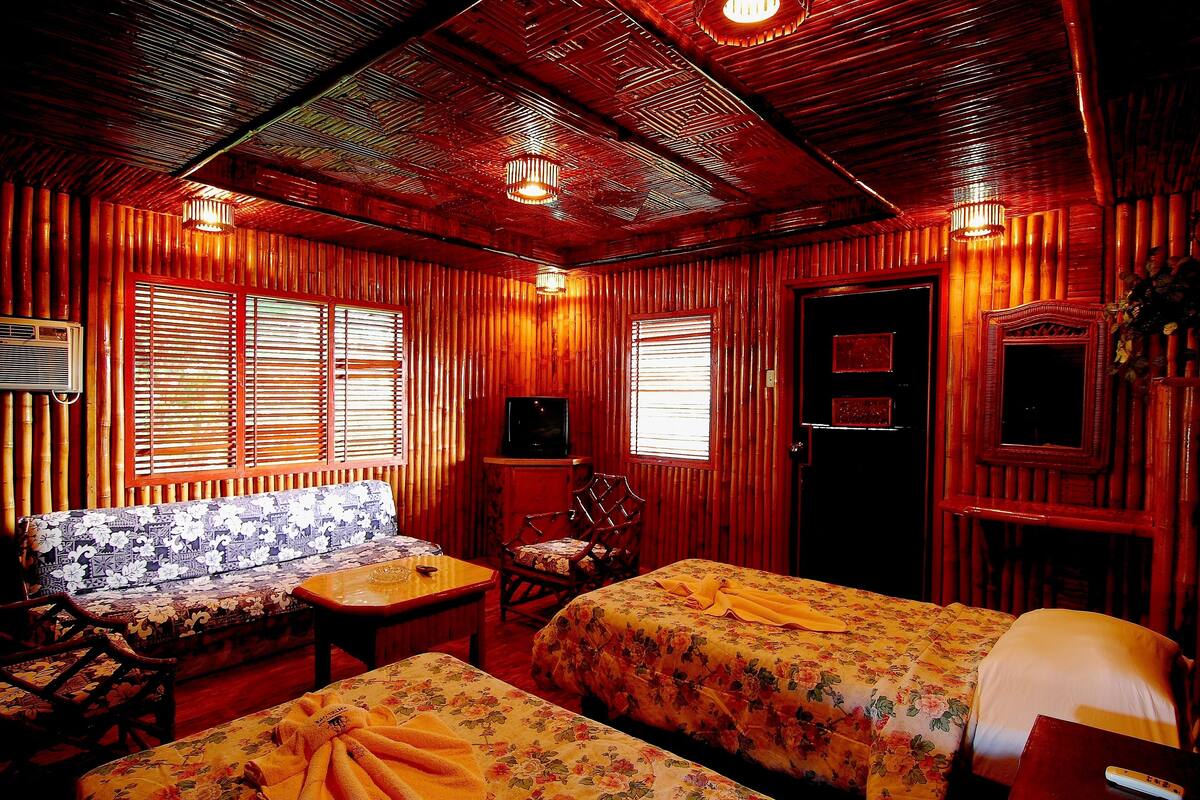 Una habitación con paredes y techo de madera, un sofá, dos camas con ropa de cama floral, una silla de madera y un televisor.