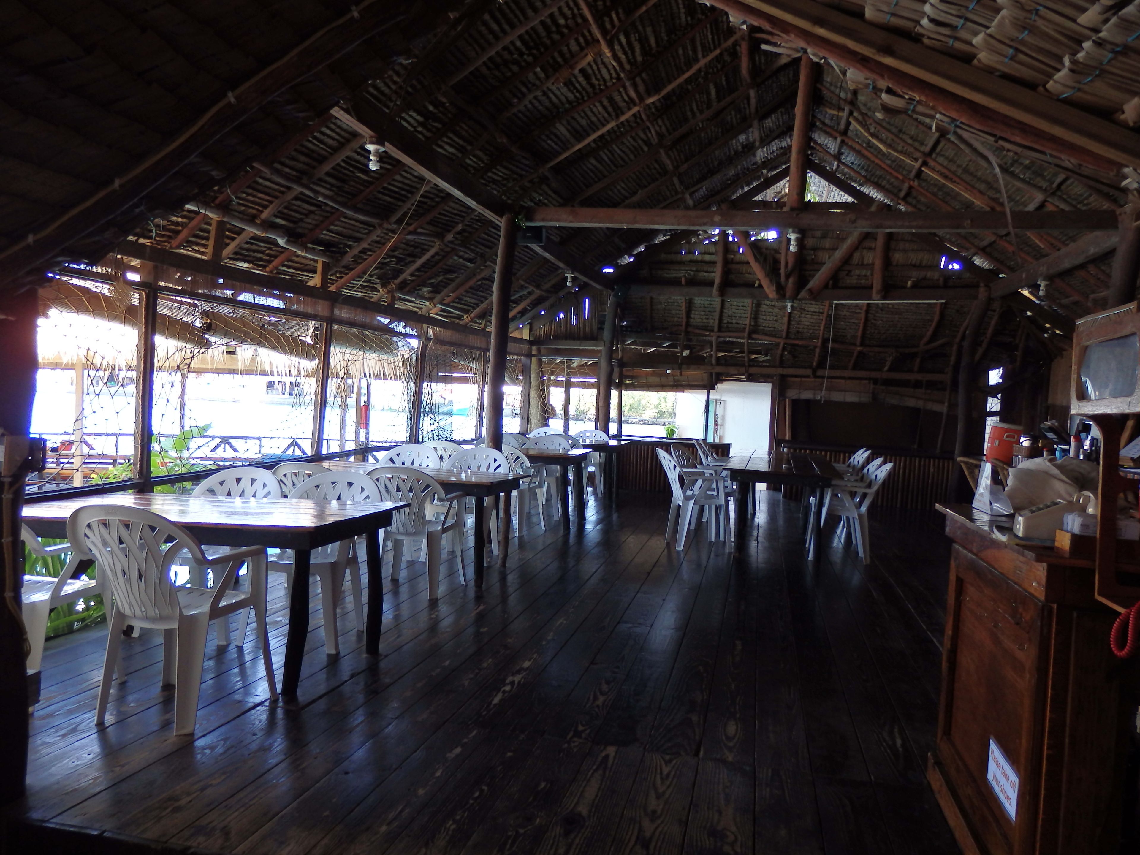 An open-air restaurant with wooden tables and white plastic chairs.