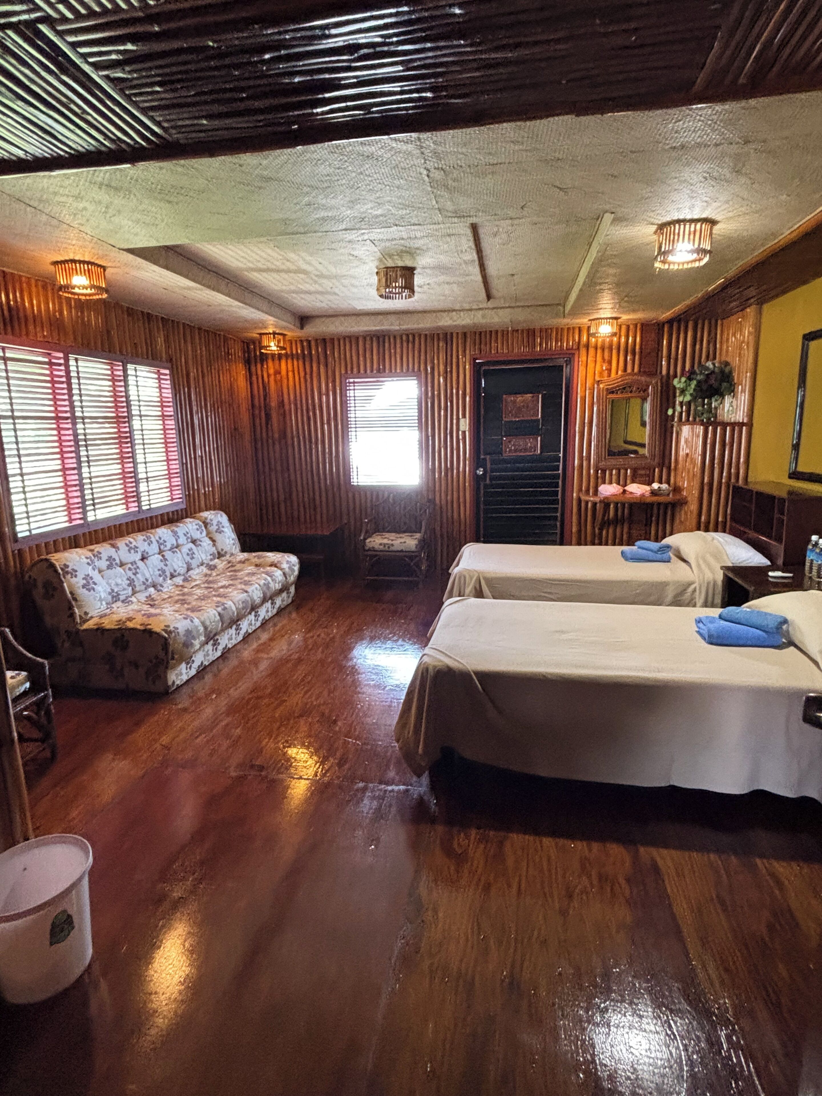 A room with wooden walls and floors, a sofa, two beds, a mirror, and a window with blinds.