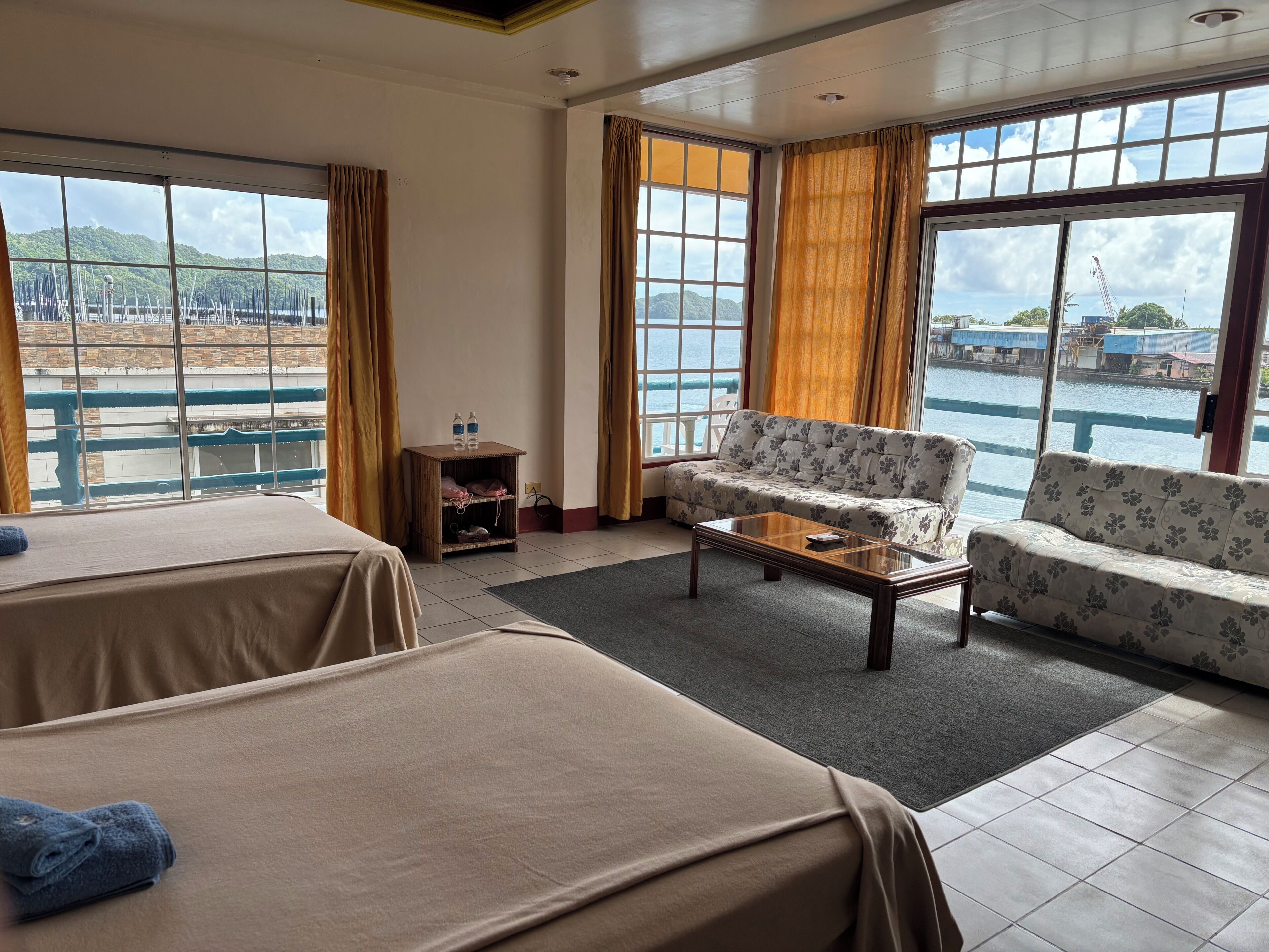 A hotel room with two beds, a sofa, a small table, and large windows offering a view of a body of water and distant hills.