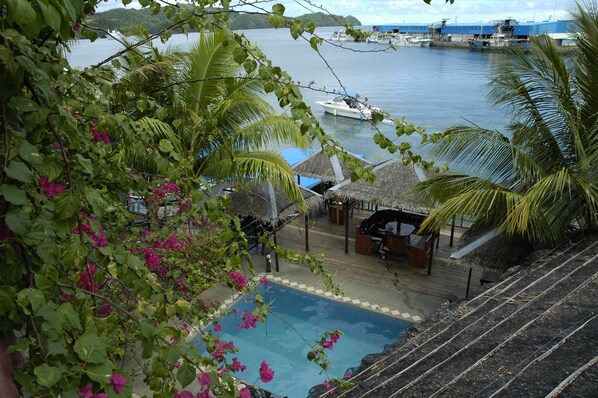 A pool area with a view of the sea, surrounded by lush greenery and a thatched-roof structure.